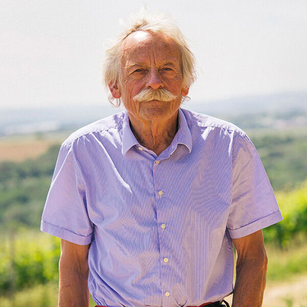 Ein Senior mit weißem Haar und Schnurrbart trägt ein hellblaues Hemd und steht vor einem malerischen Weinberg im Hintergrund.
