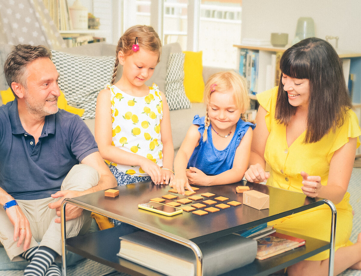 Eine Familie mit zwei Kindern spielt das Spiel 'klang² Spielesammlung »Geigenbauer«' auf einem Couchtisch. Die Szenerie zeigt ein Wohnzimmer mit viel natürlichem Licht und die Familie lächelt und genießt die Spielzeit zusammen. Das Spiel besteht aus Holzbausteinen.