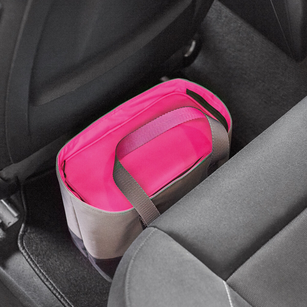 Eine pinkfarbene Lunch-Tasche befindet sich auf dem Boden des Rücksitzes in einem Auto. Die Tasche hat graue und schwarze Elemente sowie ein graues Trageband.
