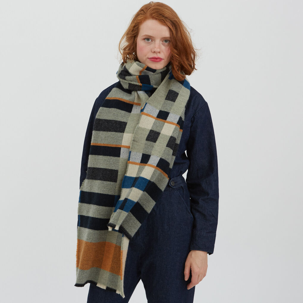 Frau mit roten Haaren trägt einen Bauhaus Stölzl Schal/Wrap. Der Schal hat ein geometrisches Muster in Grau-, Schwarz-, Braun- und Blautönen und ist um den Hals der Frau gewickelt.