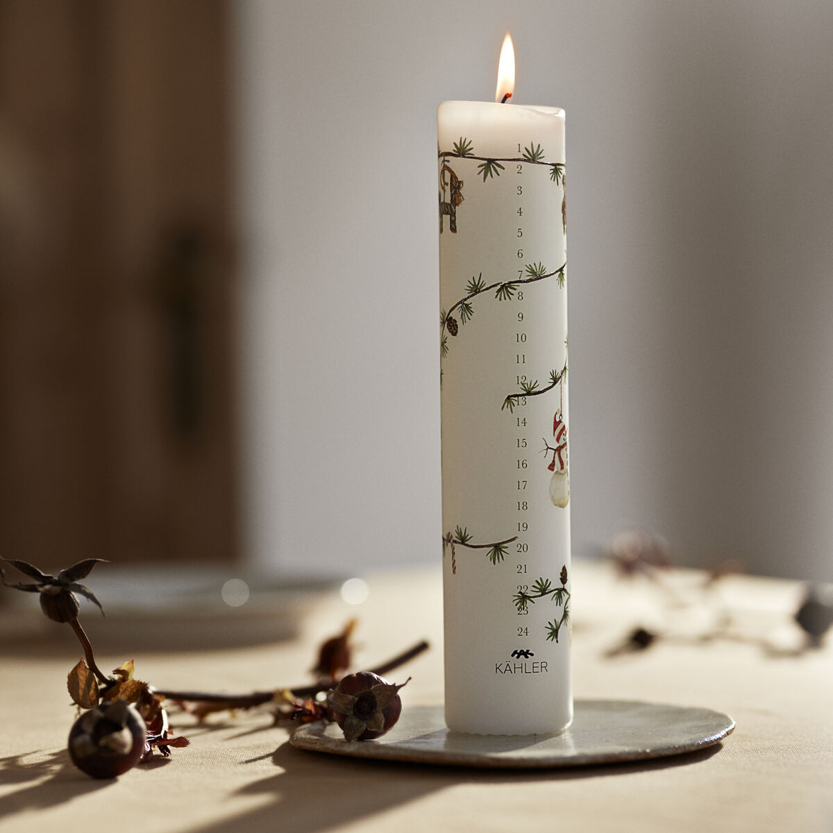 Eine weiße Adventskalenderkerze der Serie Hammershøi steht brennend auf einem runden Keramikuntersetzer auf einem gedeckten Tisch. Die Kerze ist mit Zahlen, winterlichen Zweigen und kleinen Wichtelfiguren verziert. Sonnenlicht fällt durch ein Fenster, wodurch eine warme und gemütliche Atmosphäre entsteht.