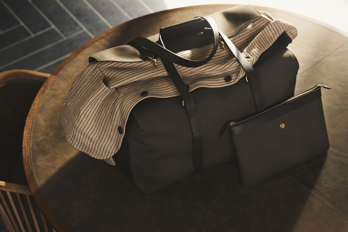 Die Abbildung zeigt eine stilvolle, große Tasche und eine kleinere Tasche namens »POUCH LARGE« auf einem Tisch. Die große Tasche ist schwarz mit Lederriemen und hat eine beige-gestreifte Textilauflage. Die kleinere Tasche ist ebenfalls schwarz mit einem goldfarbenen Reißverschluss.
