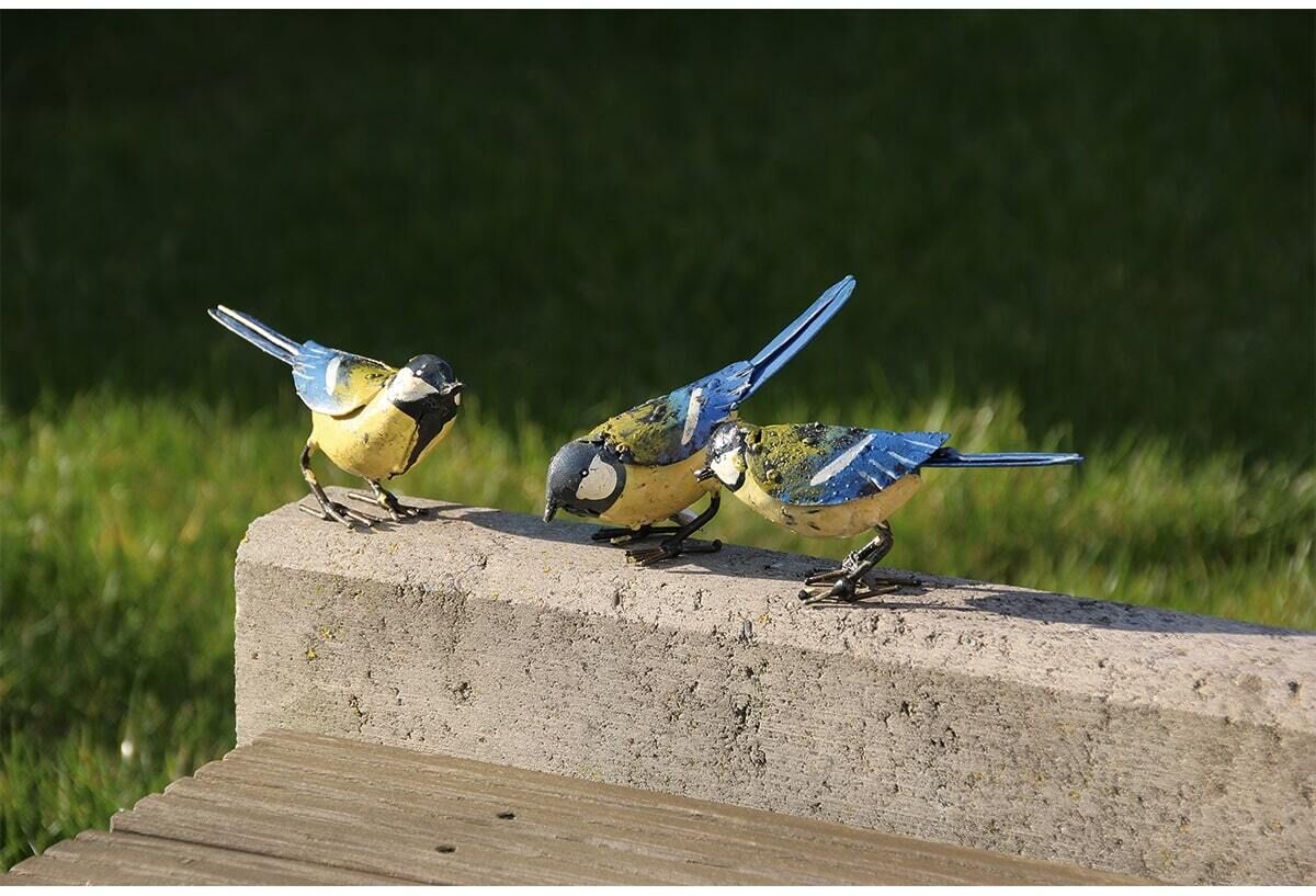 Drei dekorative Metallvögel in Blau, Grün und Gelb stehen auf einem steinernen Rand mit Gras im Hintergrund.