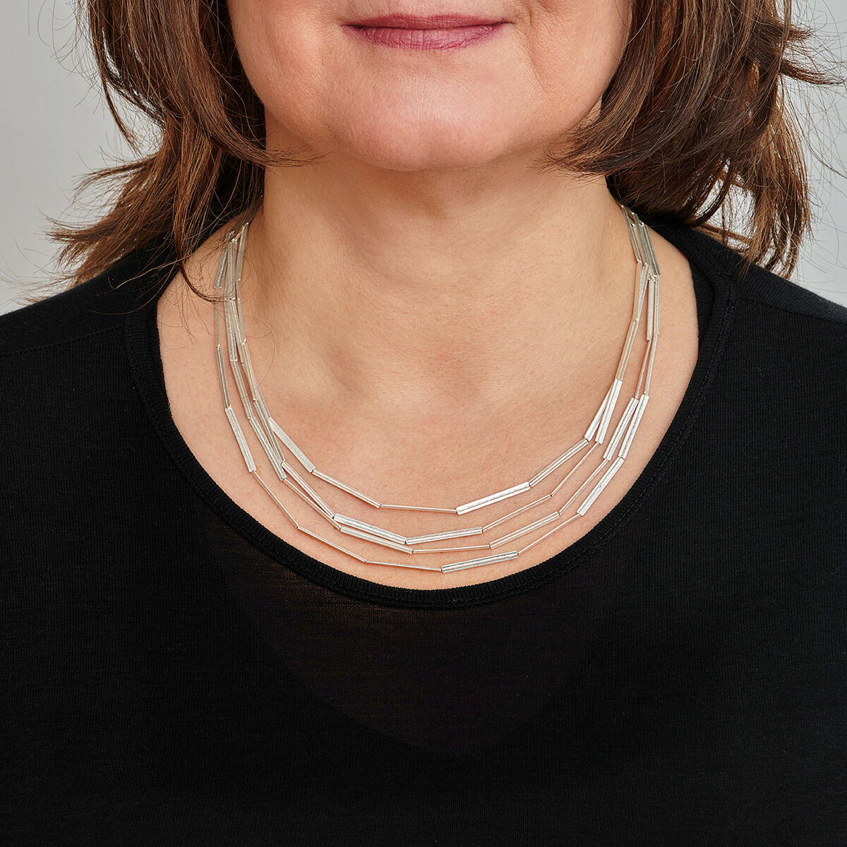 Nahaufnahme einer Frau, die eine silberne Kette mit Streifenmuster trägt. Die Kette liegt auf einem schwarzen Oberteil und besteht aus mehreren dünnen, langen Elementen.