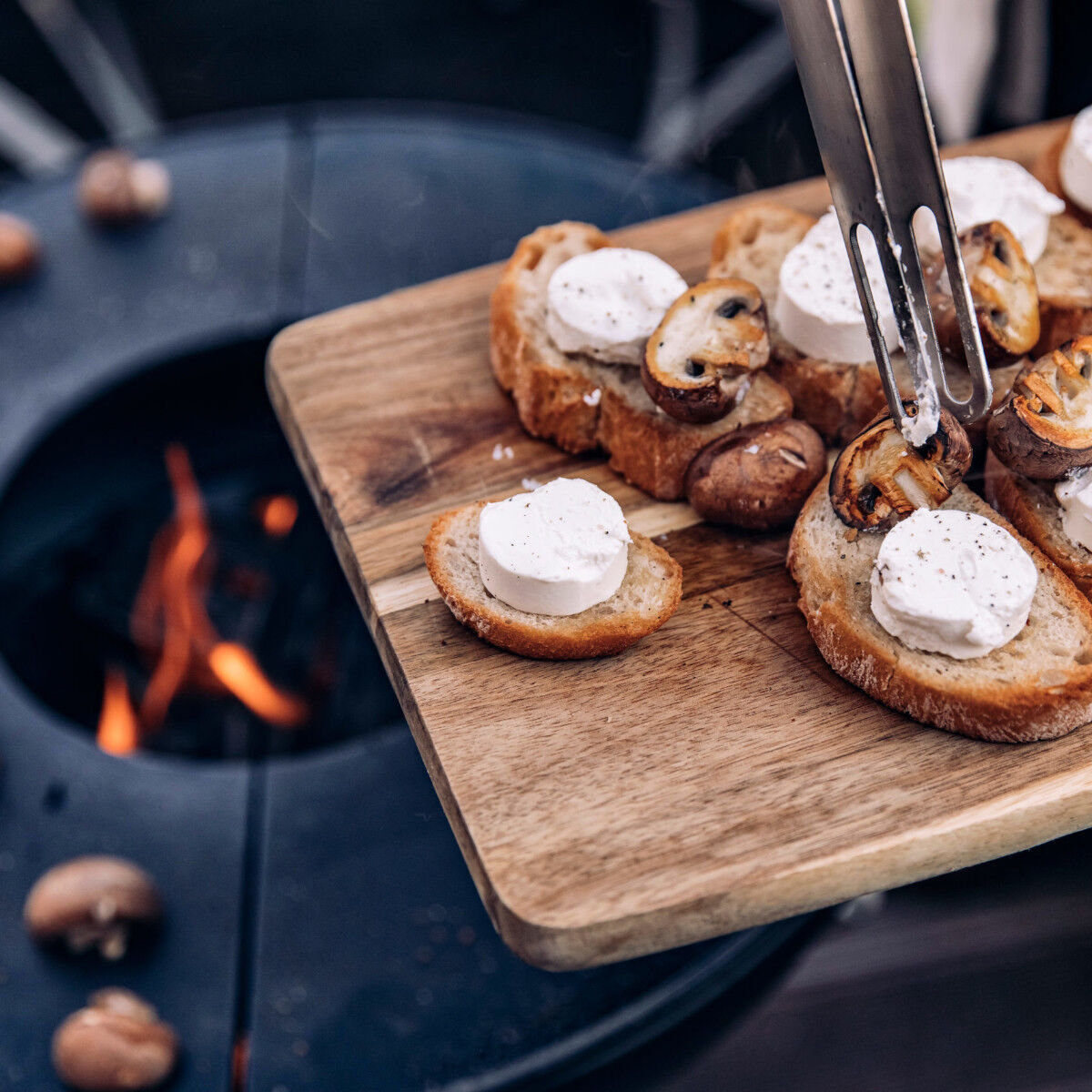 Plancha Halbring von BOWL bei der Essenszubereitung mit gegrilltem Brot und Käse auf einem Holzbrett, im Hintergrund eine Feuerstelle.