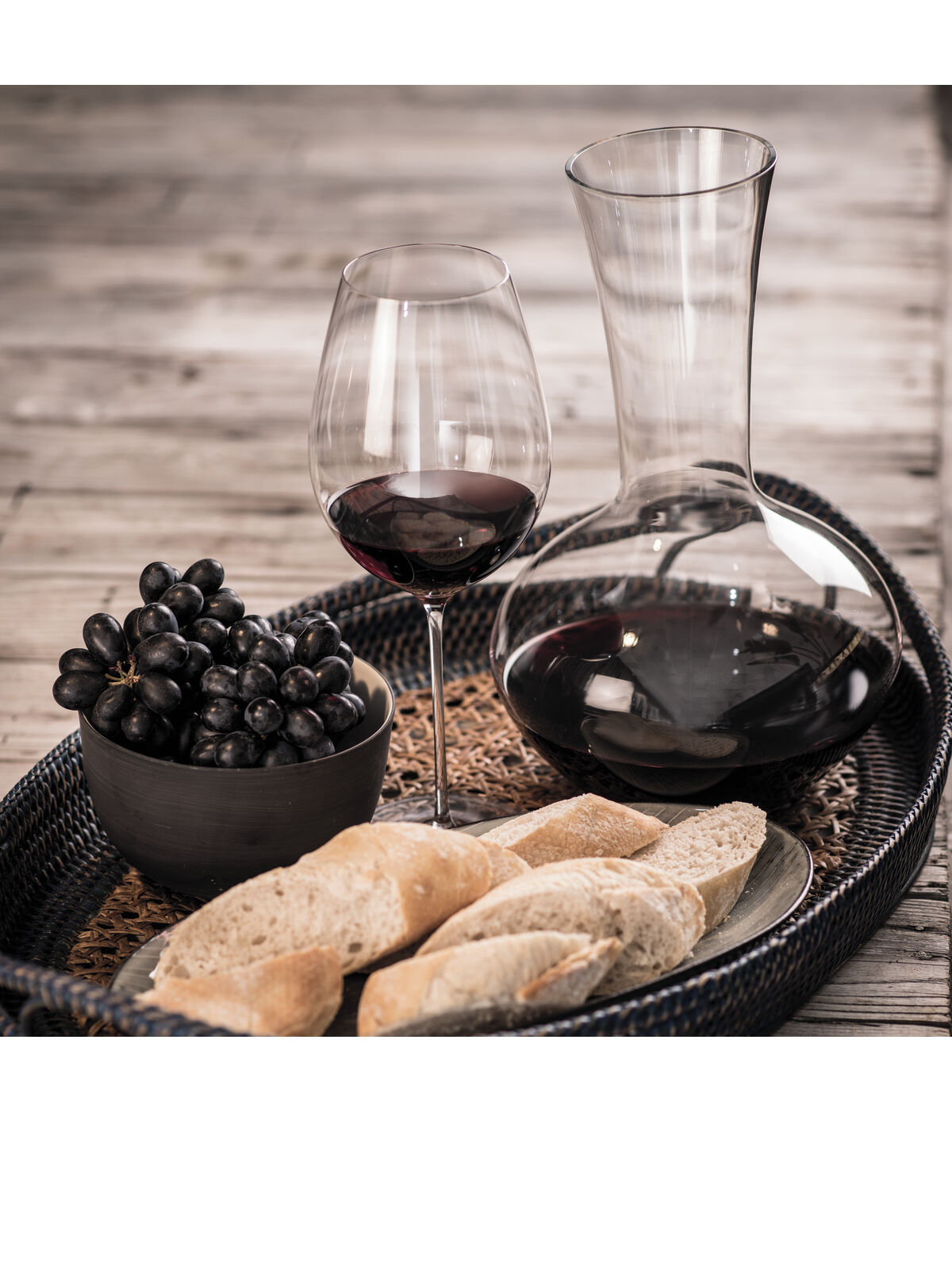 Eine Flasche Rotwein und ein Weinglas mit rotem Wein neben schwarzen Trauben in einer Schale und geschnittenem Baguette auf einem Tablett.