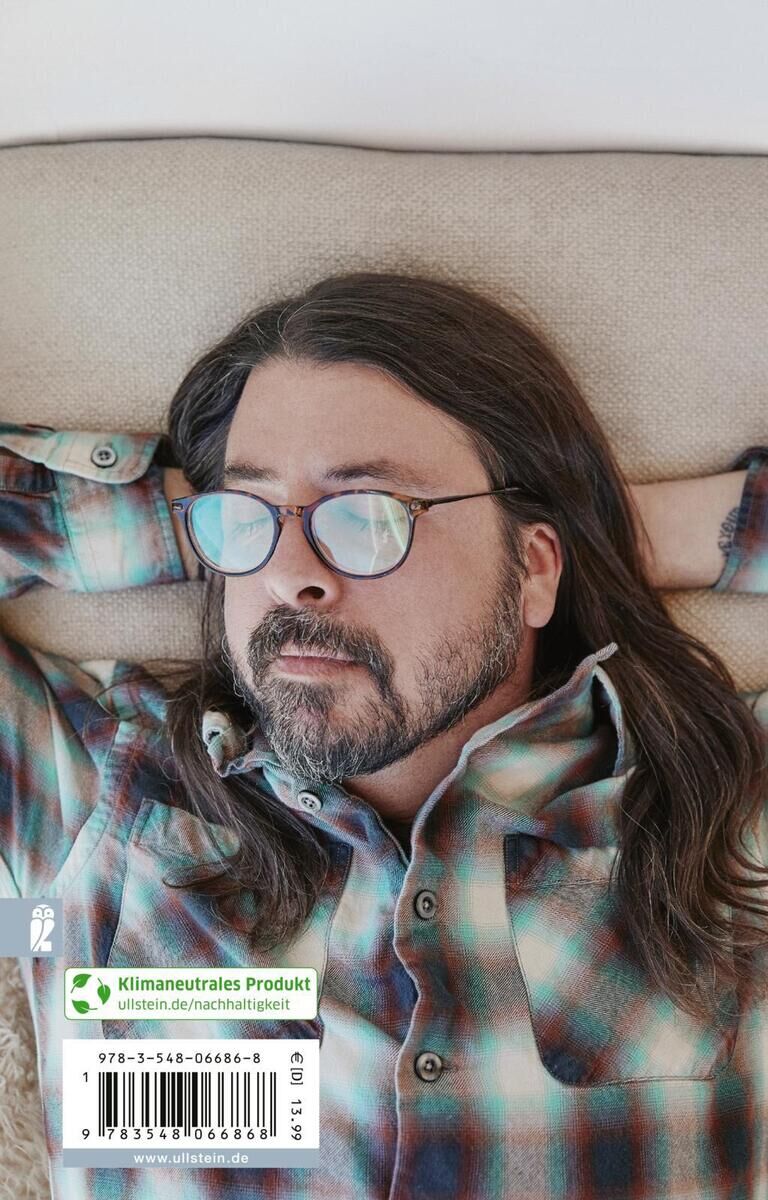 Ein Mann mit langen Haaren und einer Brille liegt entspannt auf einem beigen Sofa. Er trägt ein kariertes Hemd in Blau- und Brauntönen und hat die Arme hinter den Kopf gelegt. Auf ihm liegt ein Buch mit dem Titel 'Grohl, D: Storyteller', das ein klimaneutrales Produktsiegel und eine ISBN-Nummer zeigt.