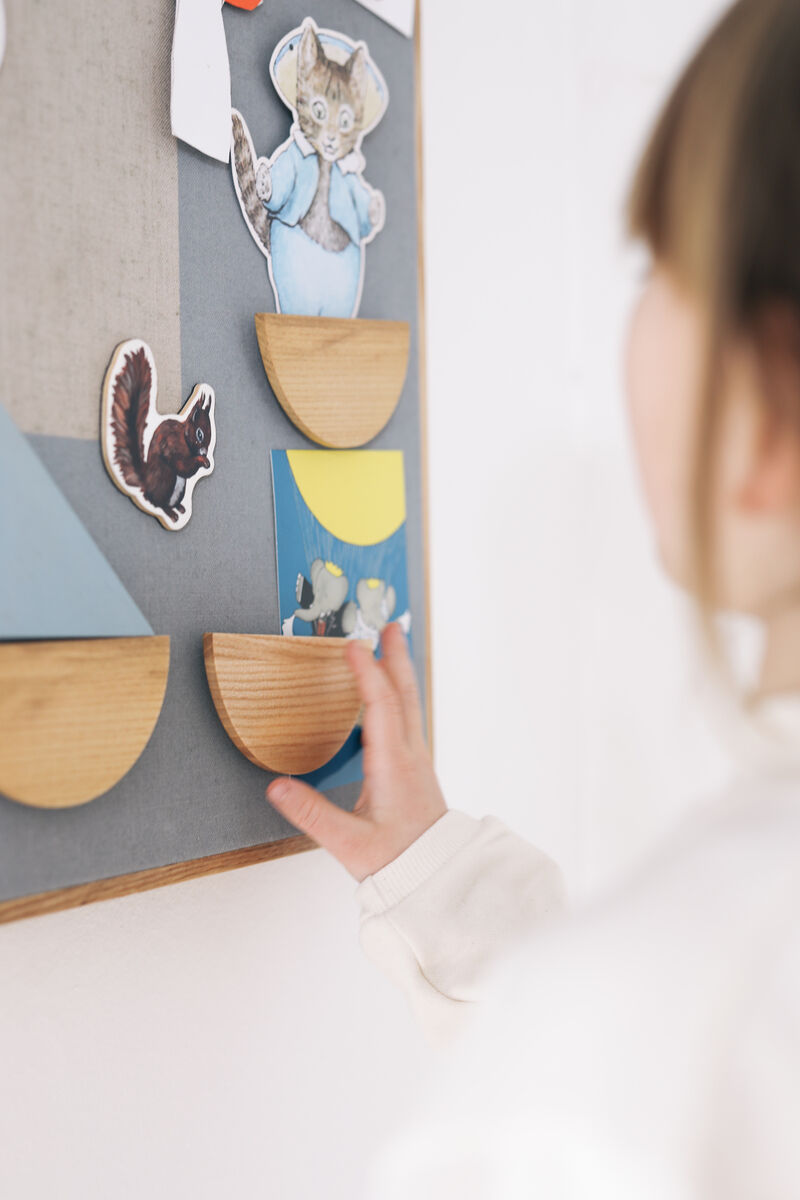 Ein Kind platziert einen Holzmagnet auf einer Pinnwand, die mit verschiedenen kindlichen und farbenfrohen Papierstücken verziert ist.