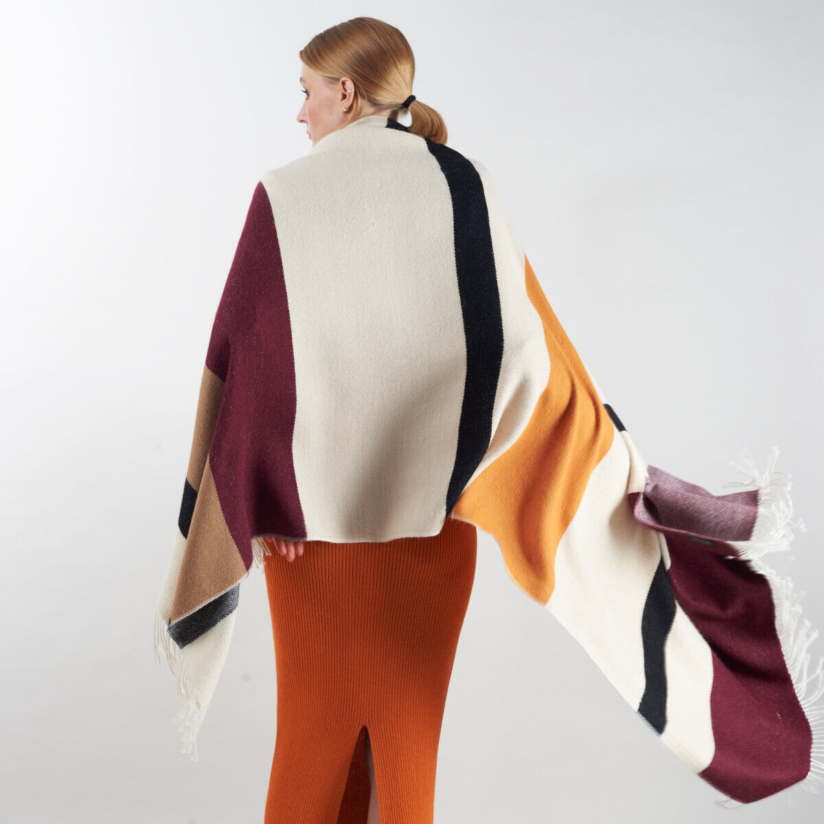 Eine Frau trägt ein großflächiges Cape »Alvar« aus Alpaka-Wolle. Das Cape zeigt ein farbenfrohes, modernes Design mit Cremeweiß, Schwarz, Bordeauxrot und Ocker, während die Frau einen orangefarbenen Strickrock trägt. Der Umhang wirkt gemütlich und stilvoll.