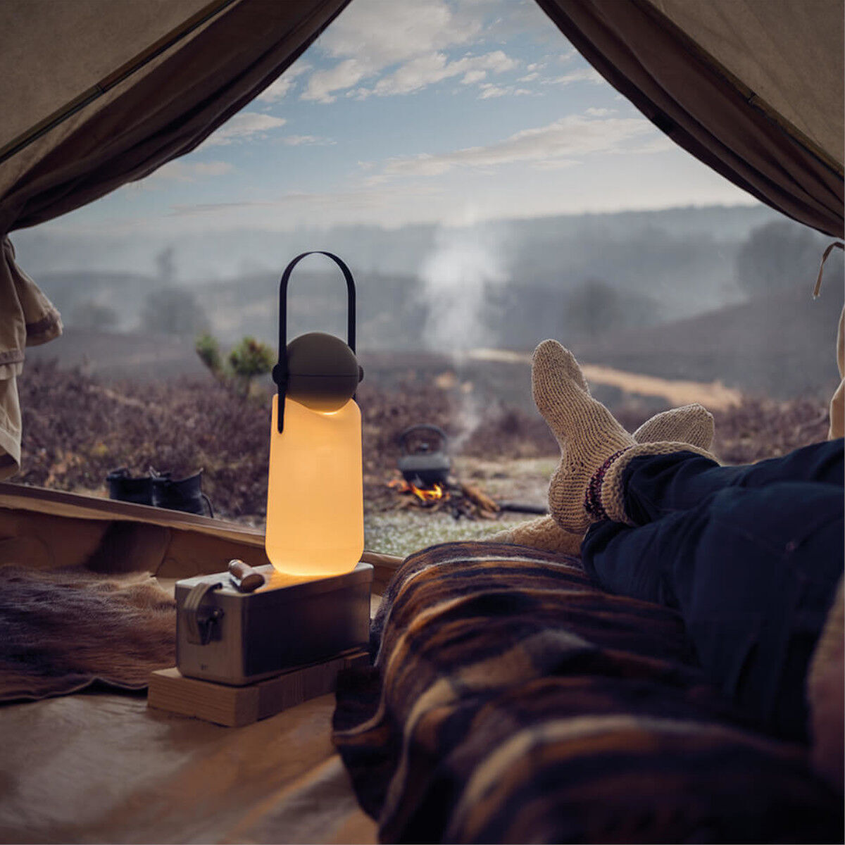 Ein stimmungsvolles Bild zeigt eine »Guidelight« Lampe auf einem Holzhocker in einem gemütlichen Zelt mit einer Aussicht auf eine neblige Berglandschaft bei Abenddämmerung. Ein Paar in Wollsocken genießt den Moment, während ein Lagerfeuer draußen brennt.