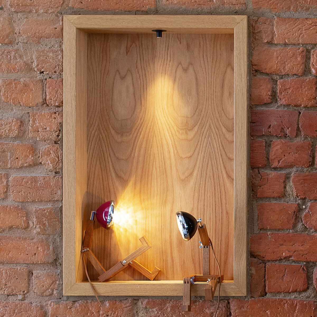 Das Bild zeigt zwei 'Mr. Wattson' Leuchten, die in Holznieschen auf einem ziegelroten Hintergrund montiert sind. Jede Leuchte besteht aus Holz und Metall und hat einen energiesparenden Scheinwerfer. Die Leuchten sitzen in den Nieschen und strahlen ein warmes Licht aus.
