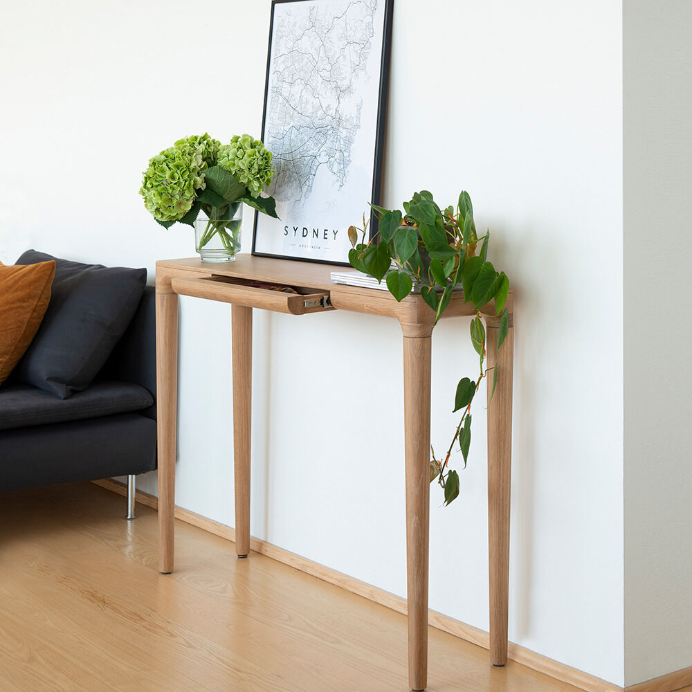 Schlanker Konsolentisch »Heart'n'Soul« aus massiver Eiche von Umage, an weißer Wand platziert. Auf der Tischplatte stehen eine Vase mit grünen Blumen, eine Pflanze und ein gerahmtes Poster. Das Setting ist hell und minimalistisch, daneben ein dunkelgraues Sofa.