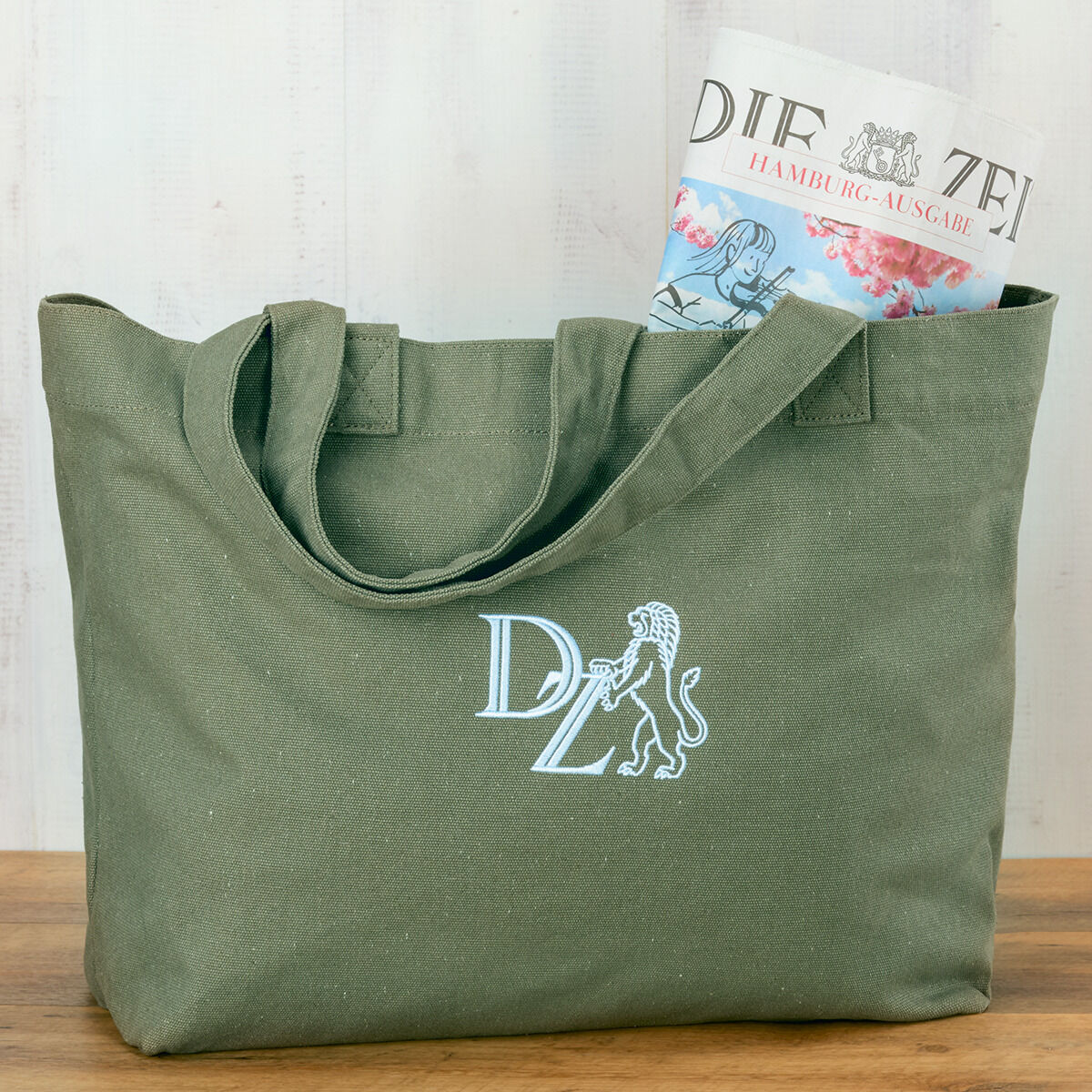 Eine olivgrüne Canvas-Tasche mit zwei Henkeln und einem stilisierten Wappenmotiv und den Buchstaben 'DZ' in Weiß. In der Tasche steckt eine ZEIT-Zeitung.