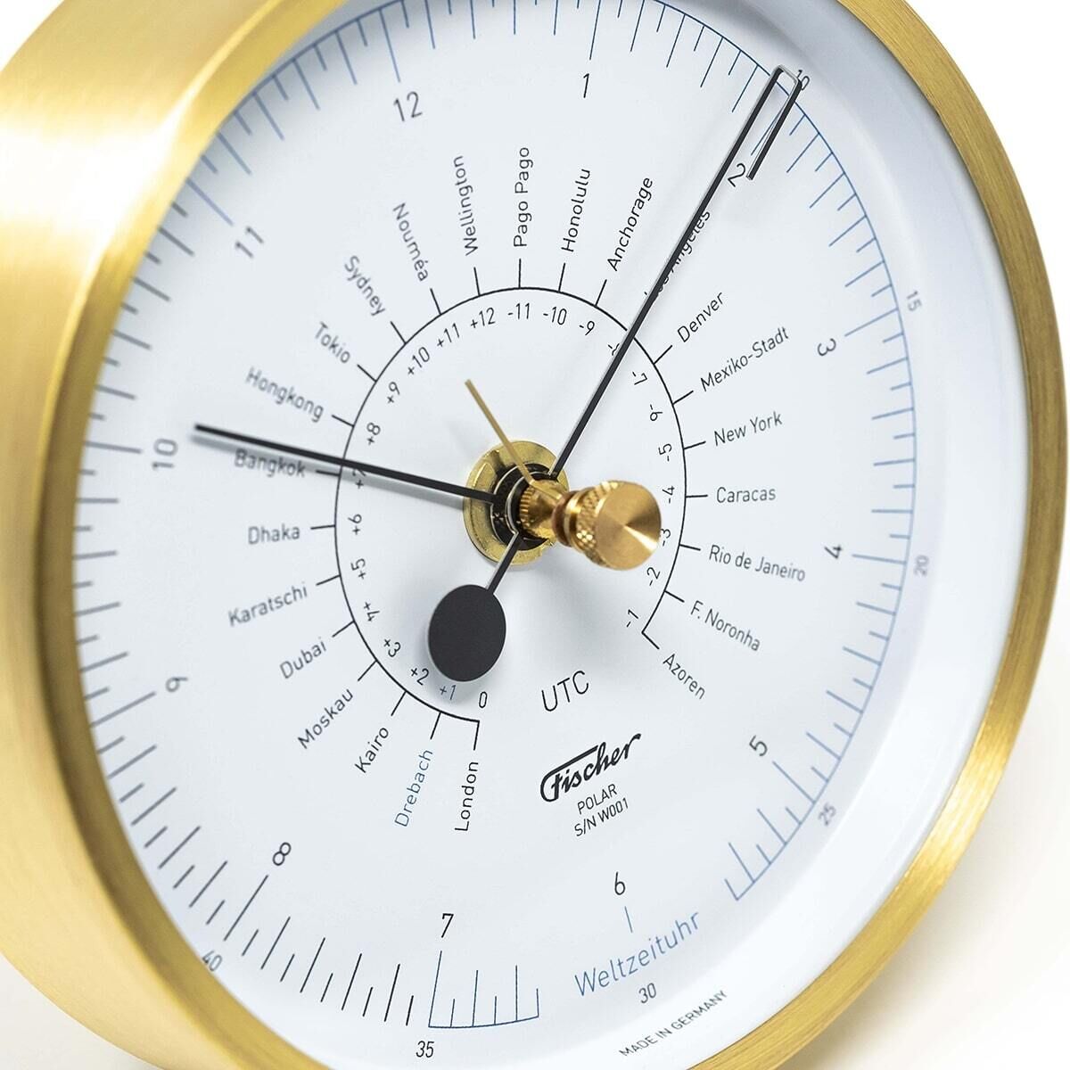 Die Weltzeituhr »POLAR« von Fischer Manufaktur ist eine moderne Uhr mit goldfarbenem Rahmen, weißem Zifferblatt und schwarzer Beschriftung, die verschiedene Zeitzonen weltweit anzeigt.