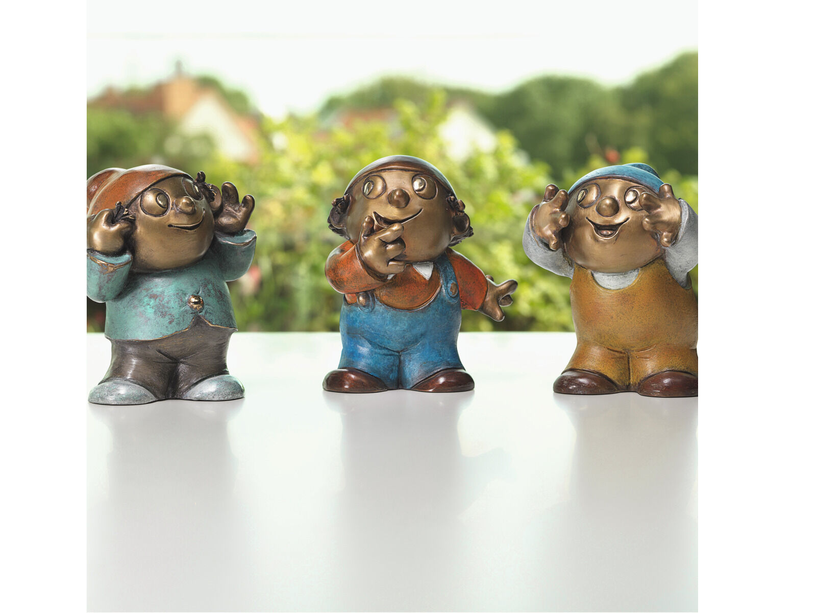Drei detailliert gearbeitete Mainzelmännchen-Skulpturen aus Bronze stehen nebeneinander auf einer glatten, weißen Fläche, im Hintergrund eine unscharfe grüne Gartenlandschaft. Jede Figur trägt unterschiedlich farbige Kleidung und zeigt individuelle, freundliche Gesichtsausdrücke und Handgesten.