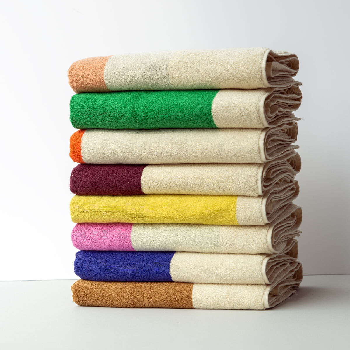 Ein Stapel von sieben Handtüchern in verschiedenen Farben (Orange, Grün, Gelb, Lila, Beige, Weiß und Braun), ordentlich aufeinander gestapelt vor einem weißen Hintergrund. Größe der Handtücher: 50 x 100 cm.