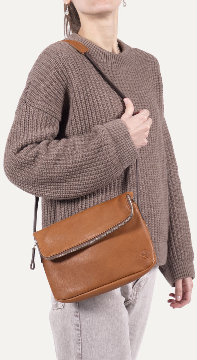 Eine Frau trägt eine mittelgroße, braune Chaza Twoway Handtasche aus glattem Kunstleder mit verstellbarem Schulterriemen. Der Crossbody-Bag passt farblich zu ihrem lockeren, beigen Strickpullover und hellgrauer Jeans. Das schlichte, moderne Design eignet sich für Alltag und Freizeit.