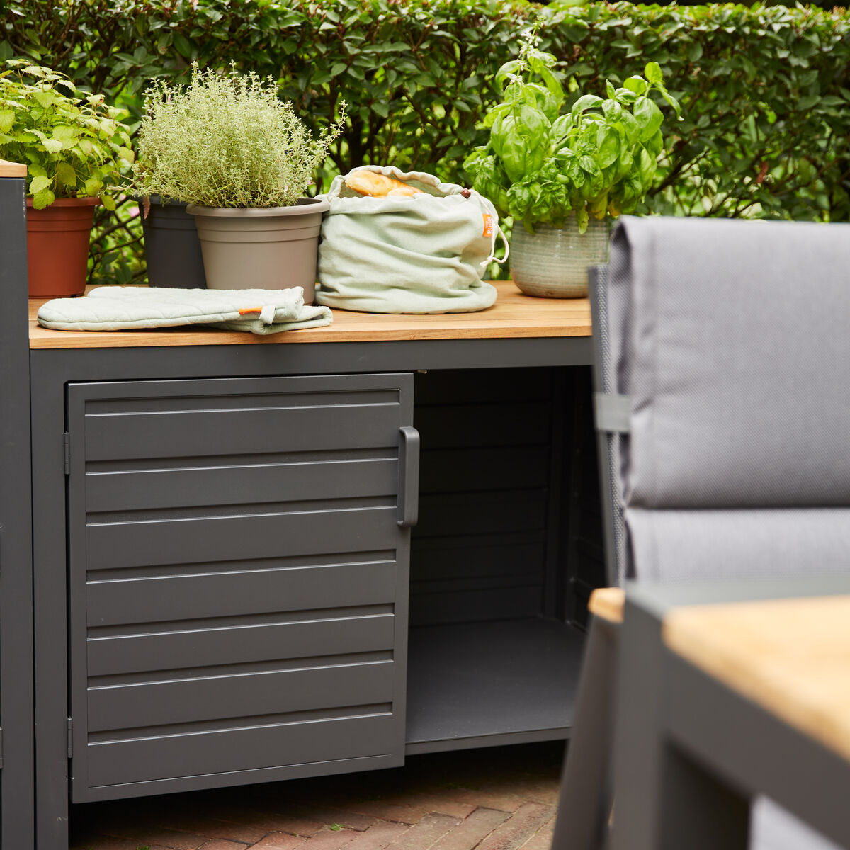 Gartenansicht einer Outdoor-Küche mit einem niedrigen Mittelteil, bestehend aus einer grauen Metallstruktur und einer Holzarbeitsplatte. Kräutertöpfe und ein Handtuch sind auf der Arbeitsfläche platziert, während im Vordergrund ein Teil eines Gartentisches und Stuhls sichtbar ist. Die Umgebung ist grün und pflanzenreich.