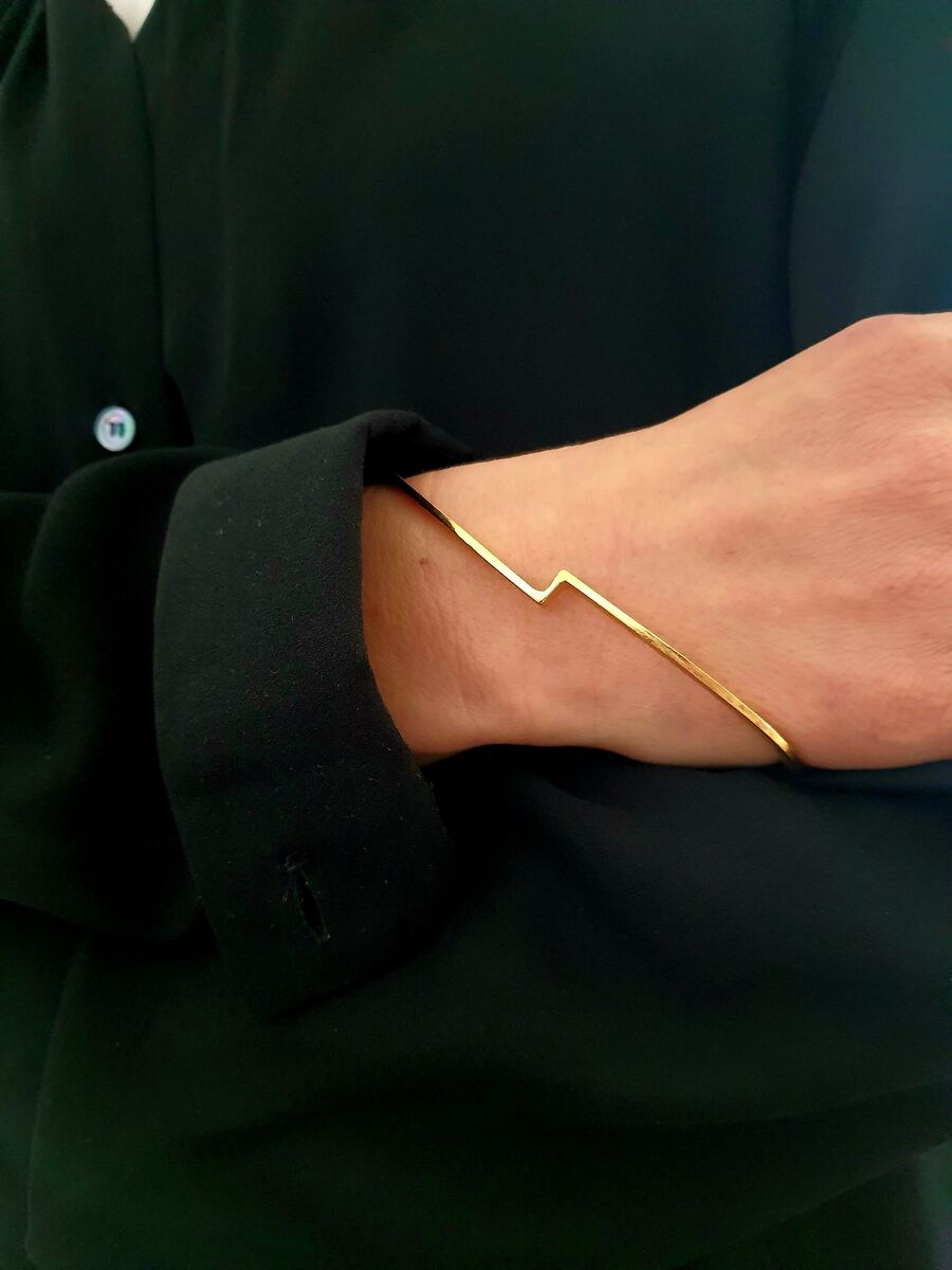 Ein goldener Armreif mit einem minimalistischen Design wird von einer Person getragen, die schwarze Kleidung anhat. Der Armreif ist schlicht und elegant.