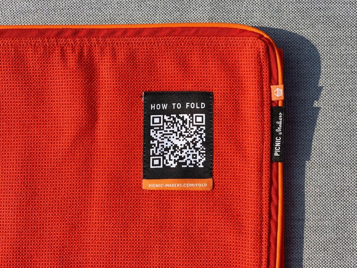 Rote Picknickdecke »Squarepark 11« mit oranger Umrandung und einem QR-Code-Label, das Anweisungen zum Falten zeigt. Hintergrund aus grauem Stoff.