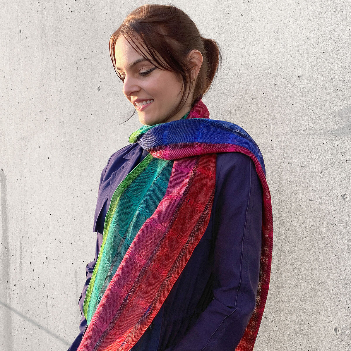 Eine Frau trägt einen buntgestreiften Leinenschal. Der Schal zeigt leuchtende Farben wie Rot, Blau, Grün und Orange. Sie trägt eine dunkelblaue Jacke und steht vor einer grauen Wand.