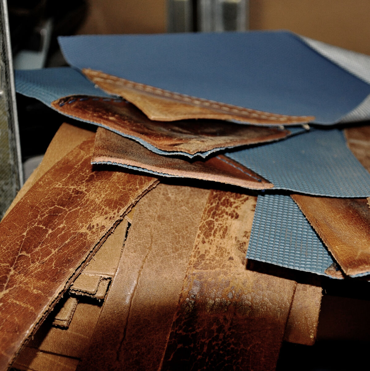 Mehrere Leder- und Stoffstücke in braunen und blauen Farbtönen liegen auf einer industriellen Oberfläche gestapelt.