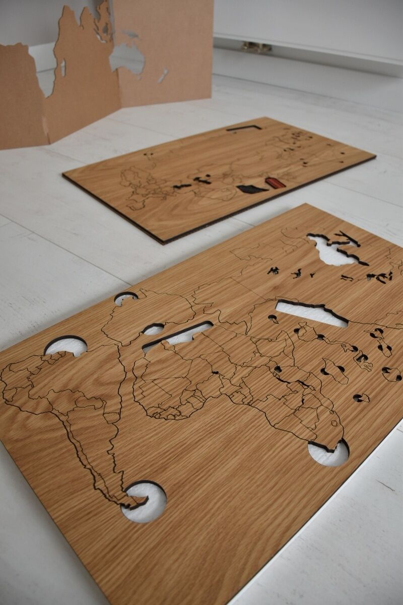 Zwei Holzbretter mit ausgeschnittenen Umrissen von Kontinenten und Ländern, die eine selbstklebende Weltkarte bilden, liegen auf einem weißen Boden. Ein Brett zeigt Umrisse Amerikas, das andere Afrika und Teile Asiens.