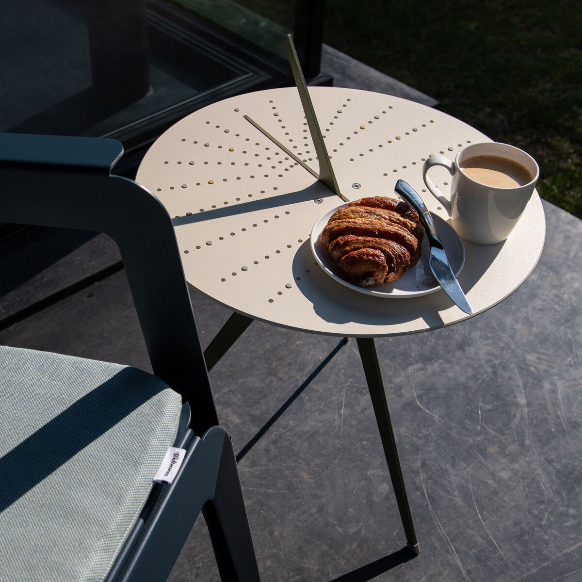 Ein runder Tisch mit Sonnenuhr-Design auf einer Terrasse, darauf eine Tasse Kaffee und ein Croissant auf einem Teller. Daneben ein Stuhl in der Sonne.