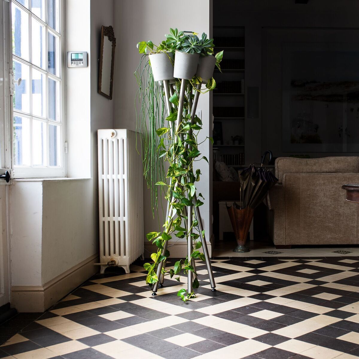 Ein stilvoller Hochgarten mit grünen Zimmerpflanzen auf einem modernen, eleganten Pflanzenständer steht in einem hellen Wohnzimmer mit schwarz-weiß gemustertem Boden und beigen Wänden.