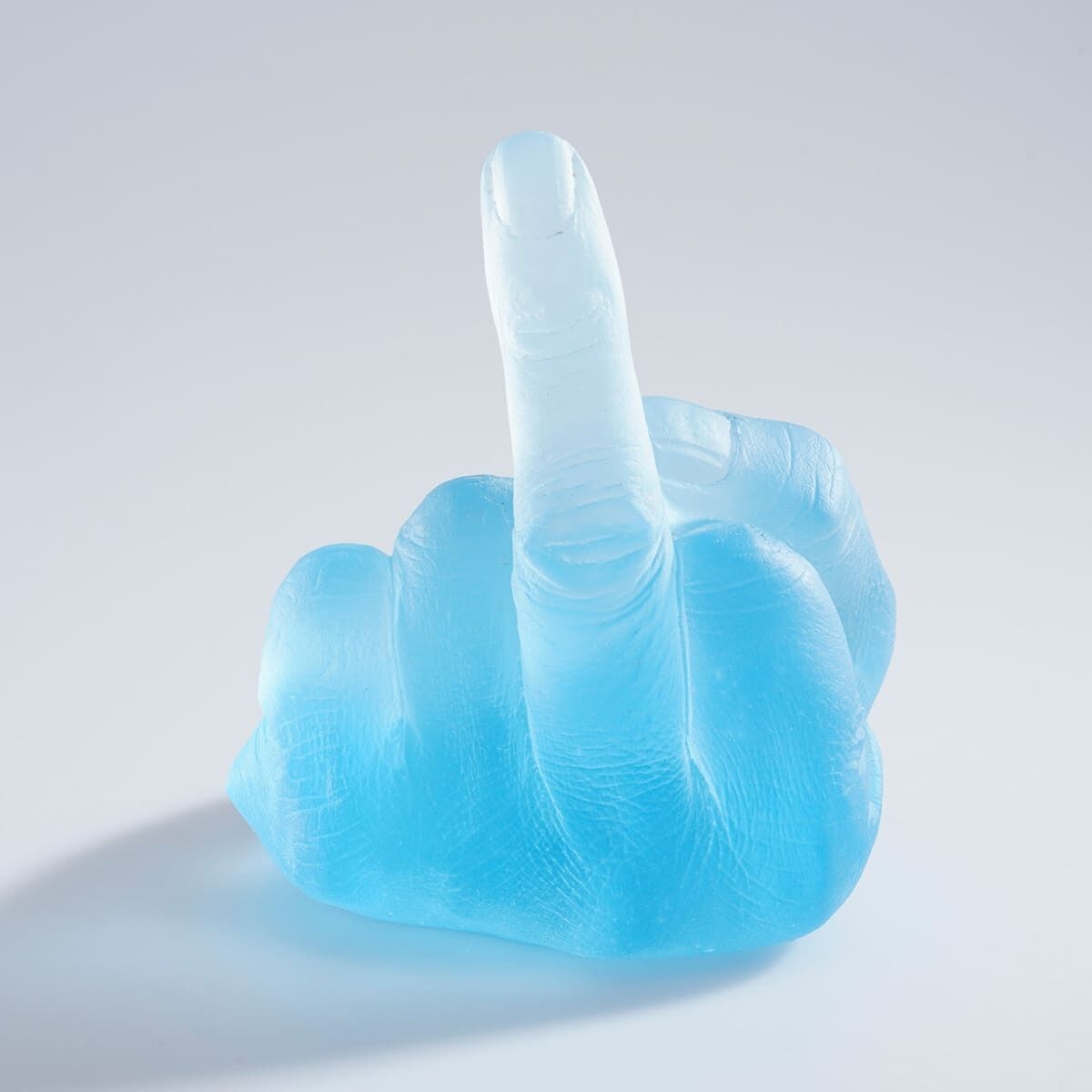 Eine Skulptur aus bläulich-weißem Glas, die eine erhobene Mittelgeste zeigt. Das Kunstwerk vereint moderne und provokative Elemente.