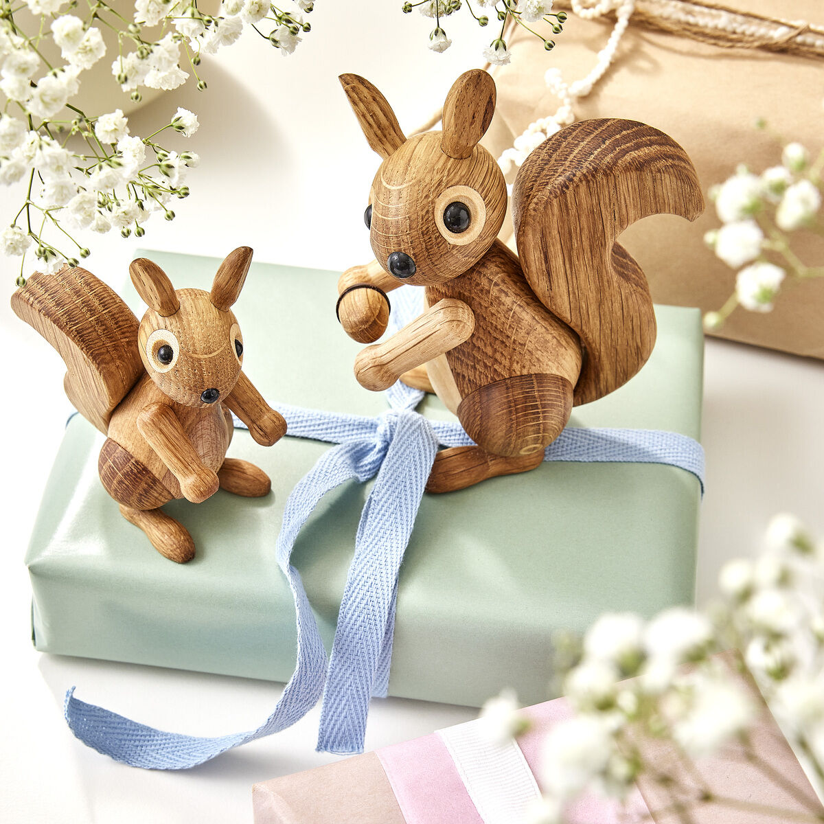 Zwei detaillierte Eichhörnchen-Holzfiguren mit natürlichen Holzmaserungen stehen auf pastellfarben verpackten Geschenken, umgeben von weißen Blumen und Dekoration, ideal als Geschenk oder Dekoration für verschiedene Anlässe.
