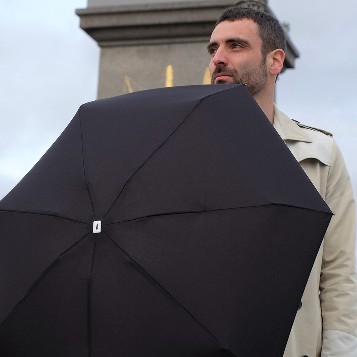 Ein Mann hält einen Taschenschirm in Schwarz. Er trägt einen beigen Trenchcoat. Im Hintergrund sind Teile einer Stadt zu sehen.