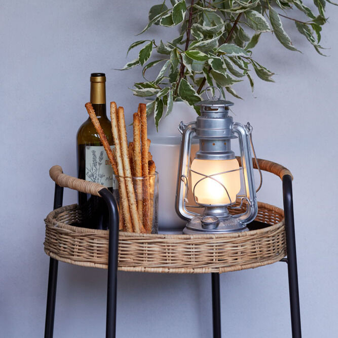 Ein Rattantisch mit einer brennenden LED-Laterne »Baby Special 276«, einer Weinflasche, Grissini-Stangen in einem Behälter, und einer Vase mit grünem Laub. Im Hintergrund hängt eine Wanddekoration.
