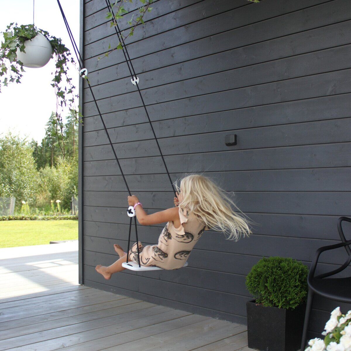 Ein Kind schaukelt auf einer modernen Kinderschaukel »Classic« an einer Terrasse mit dunkler Holzverkleidung. Im Hintergrund befindet sich ein grüner Garten und verschiedene Pflanzen. Die Schaukel ist als sicheres Outdoor-Spielgerät gestaltet und bringt Spaß an der frischen Luft.