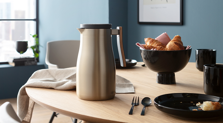 Edelstahl-Thermoskanne auf rundem Holztisch mit schwarzer Schale voller Croissants, schwarzem Geschirr und Besteck vor blauem Wandhintergrund