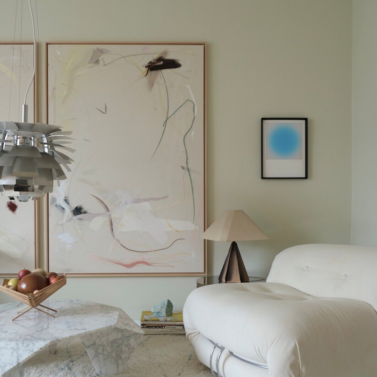 Modern eingerichtetes Wohnzimmer mit hellem, cremefarbenem Teppich und Loungesessel. An der wandgroße abstrakte Kunstwerke, darunter der Fine-Art-Print »Sorbet« von Studio Anastasia Benko, gerahmt in Holz. Auf dem Marmortisch stehen Früchte, daneben eine Designleuchte und eine stilvolle Tischlampe.