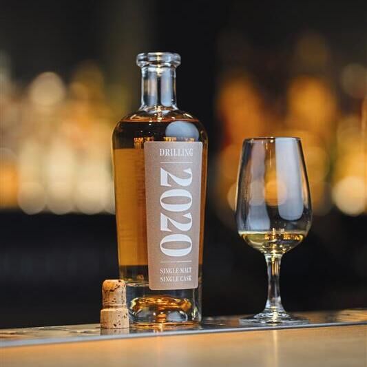 Eine stilvolle Flasche ZEIT Sonderedition Drilling Single Malt Whiskey Jahrgang 2020 steht neben einem halb gefüllten Whiskeyglas auf einer Bar. Im Hintergrund erscheinen unscharfe, warme Lichter. Der Korken der Flasche liegt auf dem Bartresen.
