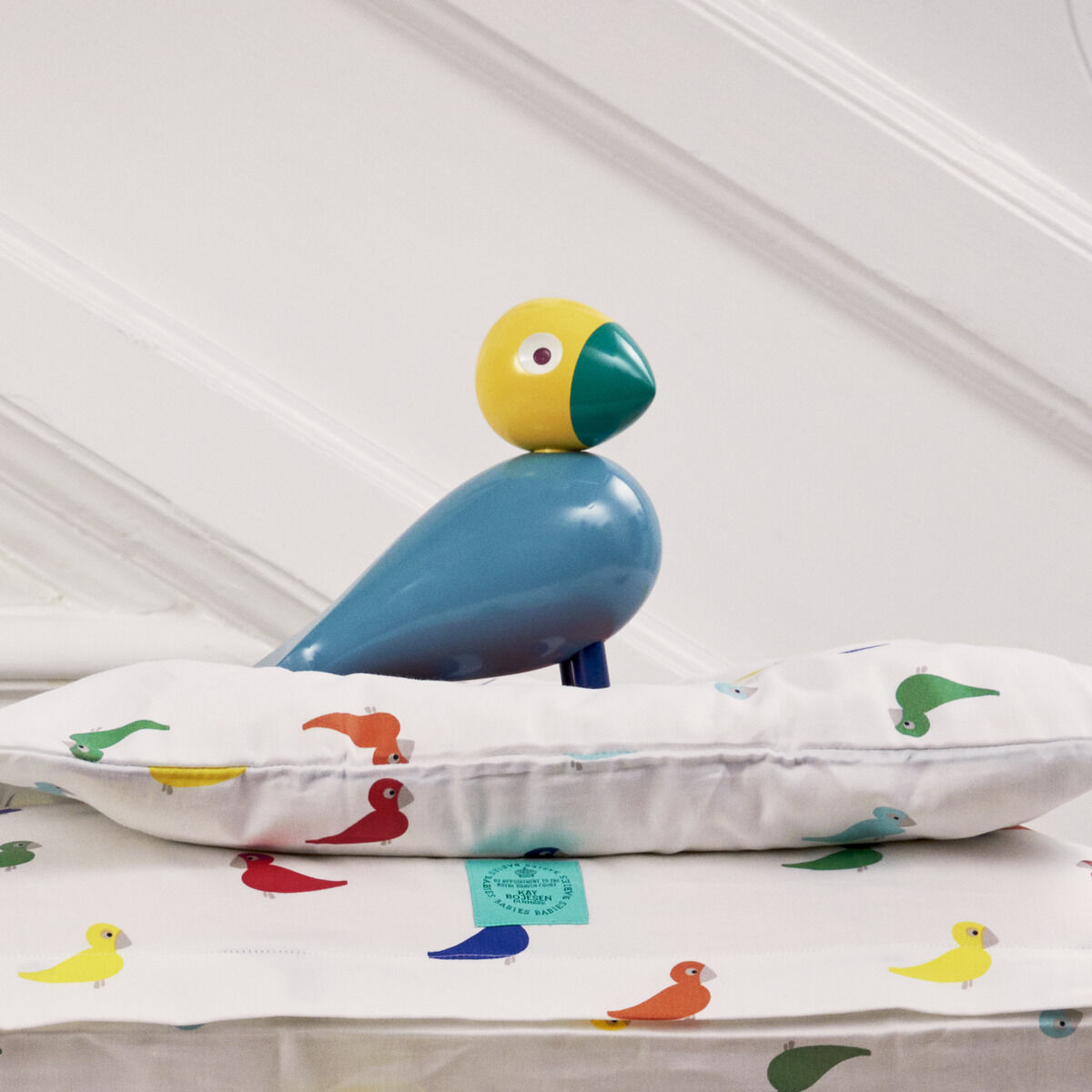 Ein Singvogel 'Sunshine' von Kay Bojesen sitzt auf einem Stapel von bunten Kissen auf einer Holzbank vor einer weißen Wand mit Holzboden.