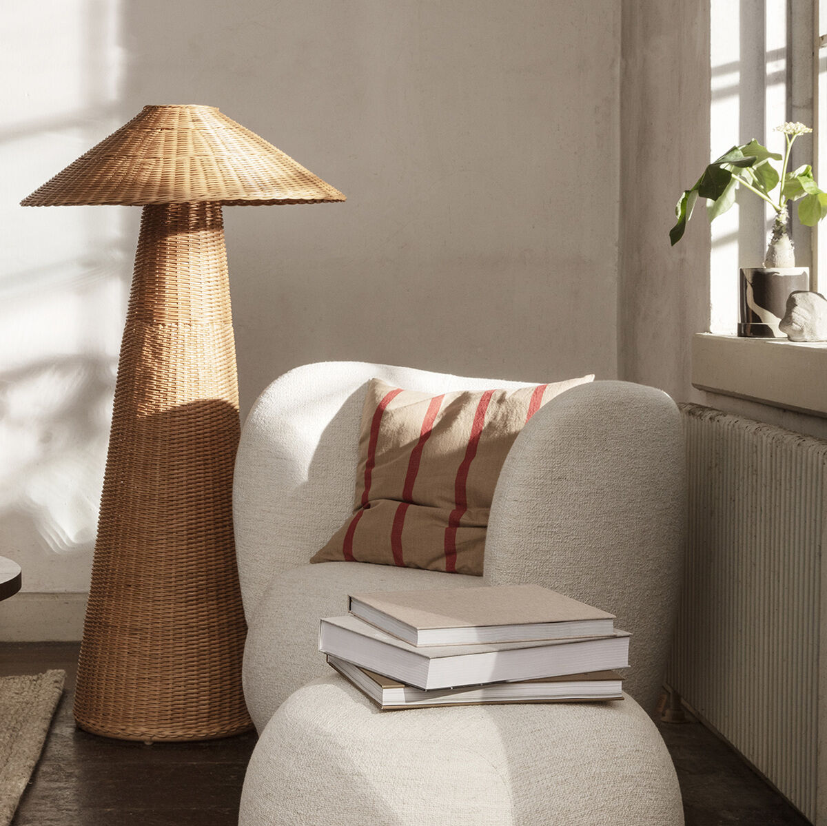Skandinavisches Wohnzimmer mit modernem, gemütlichem Sessel und Hocker in Weiß. Ein gestreiftes Kissen aus Baumwolle und Leinen rundet das Bild ab. Eine Rattan-Stehlampe und ein kleiner Tisch mit Büchern stehen in der Ecke, während ein Fenster im Hintergrund natürlichen Lichteinfall bietet.