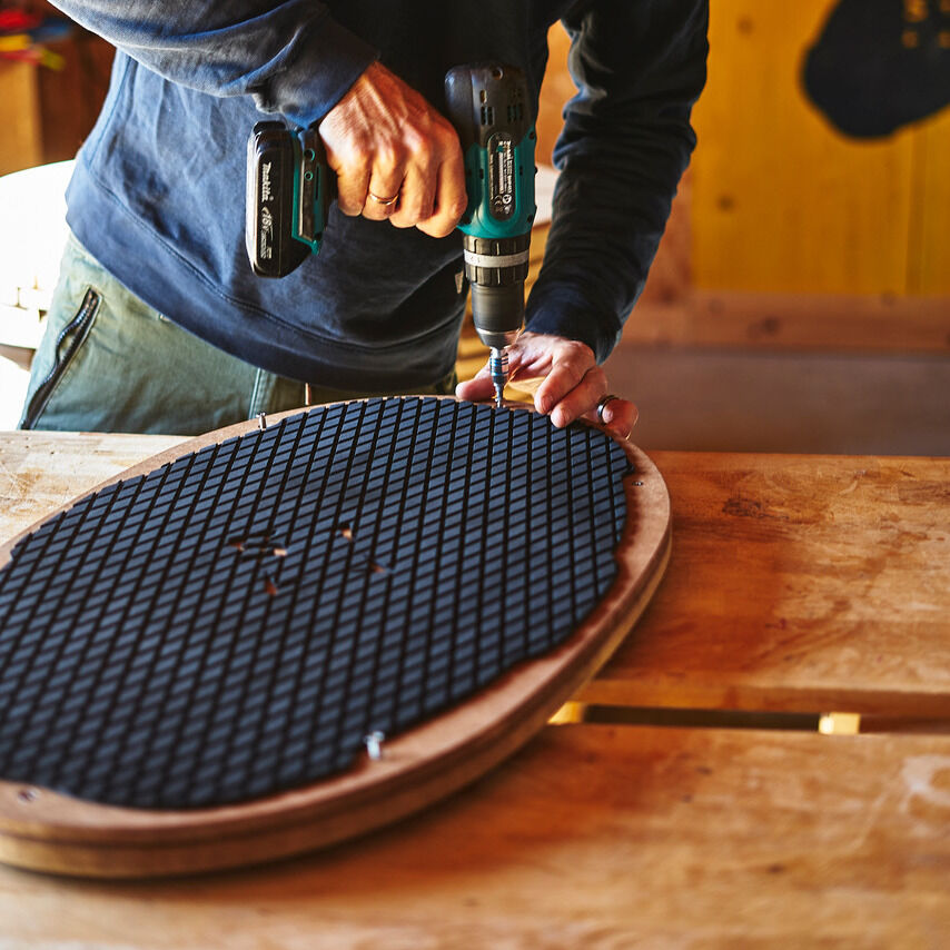 Person in blauem Pullover benutzt eine Bohrmaschine, um ein Sensoboard aus Holz mit schwarzer rutschfester Oberfläche auf einer Holzwerkbank zu befestigen.