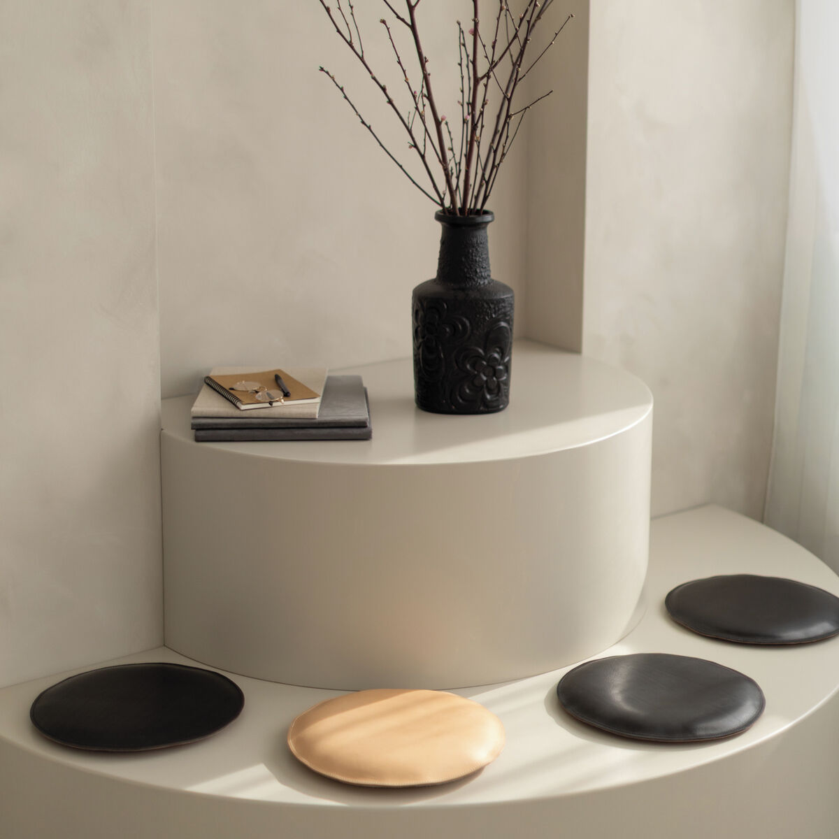 Auf einer modernen, runden Sitzfläche mit cremefarbenem Finish liegen mehrere runde Sitzpolster, davon überwiegend schwarz und eines beige. Im Hintergrund steht eine schwarze Vase mit kahlen Zweigen sowie einige Bücher als Dekoration.