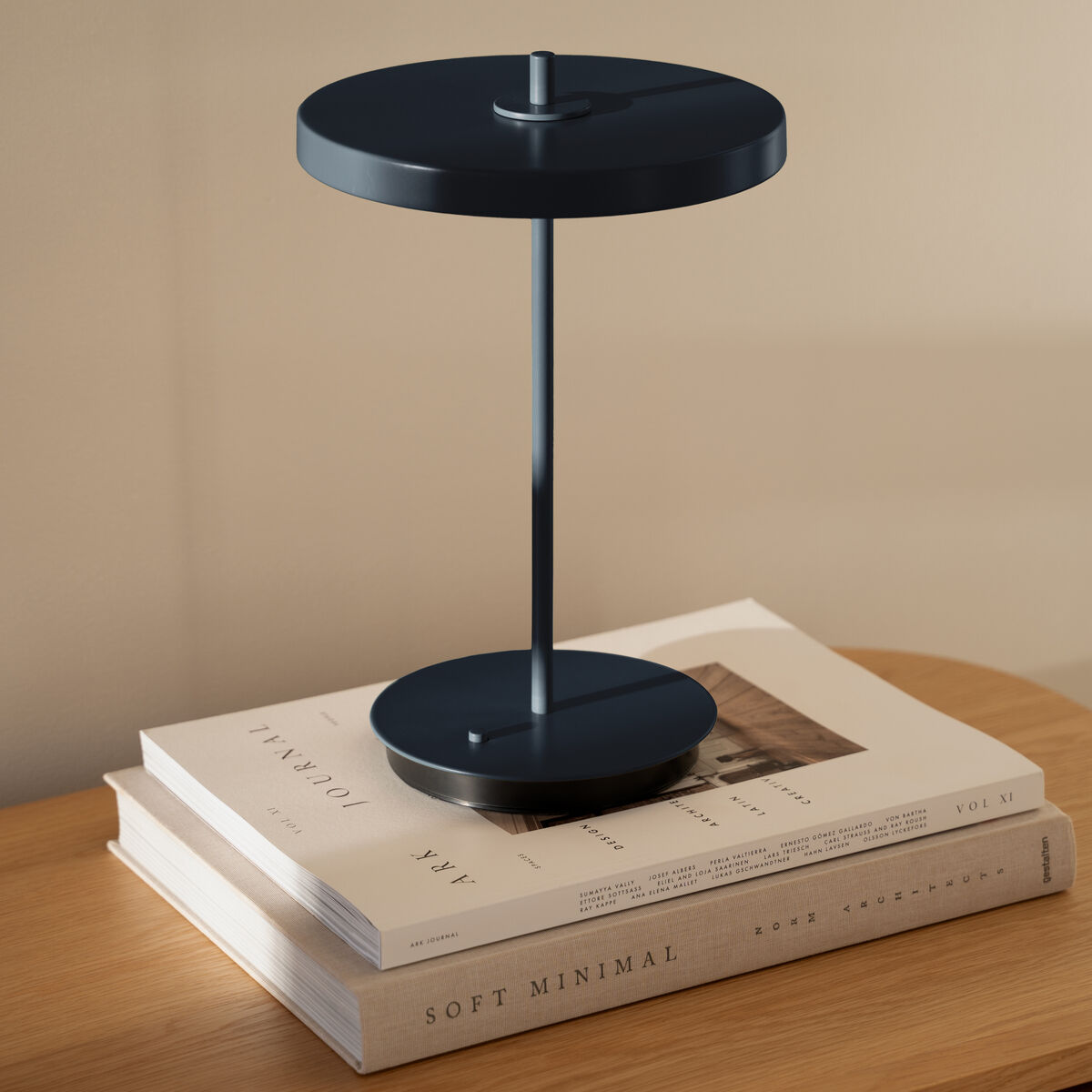 Eine moderne Tischlampe in Mitternachtsblau aus der ZEIT Sonderedition Asteria Move steht auf zwei gestapelten Büchern auf einer Holzkommode. Das minimalistische Design betont klare Linien und einen eleganten skandinavischen Stil.