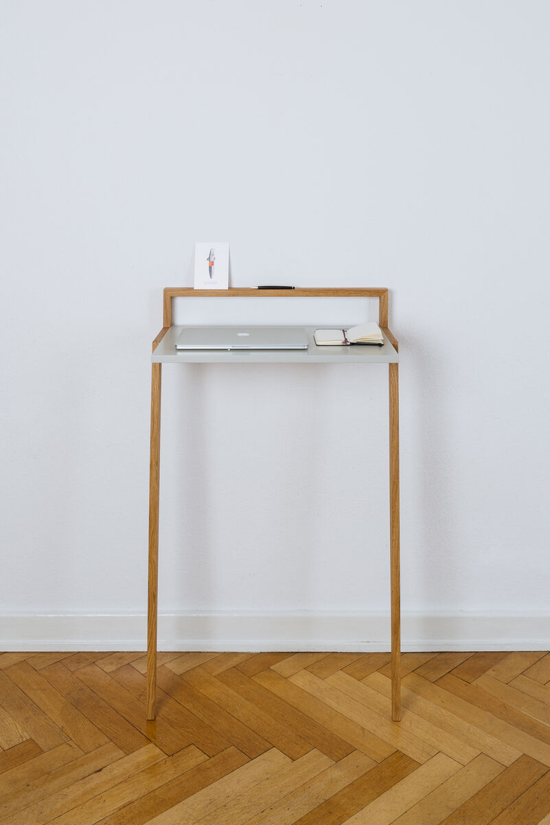Ein minimalistisches Stehpult »buckowski« aus hellem Holz, das sich ideal für das Homeoffice oder als moderner Arbeitsplatz eignet. Auf der oberen Ablagefläche sind ein kleiner Laptop und ein Notizbuch platziert. Der Pult steht vor einer weißen Wand auf einem Holzboden.