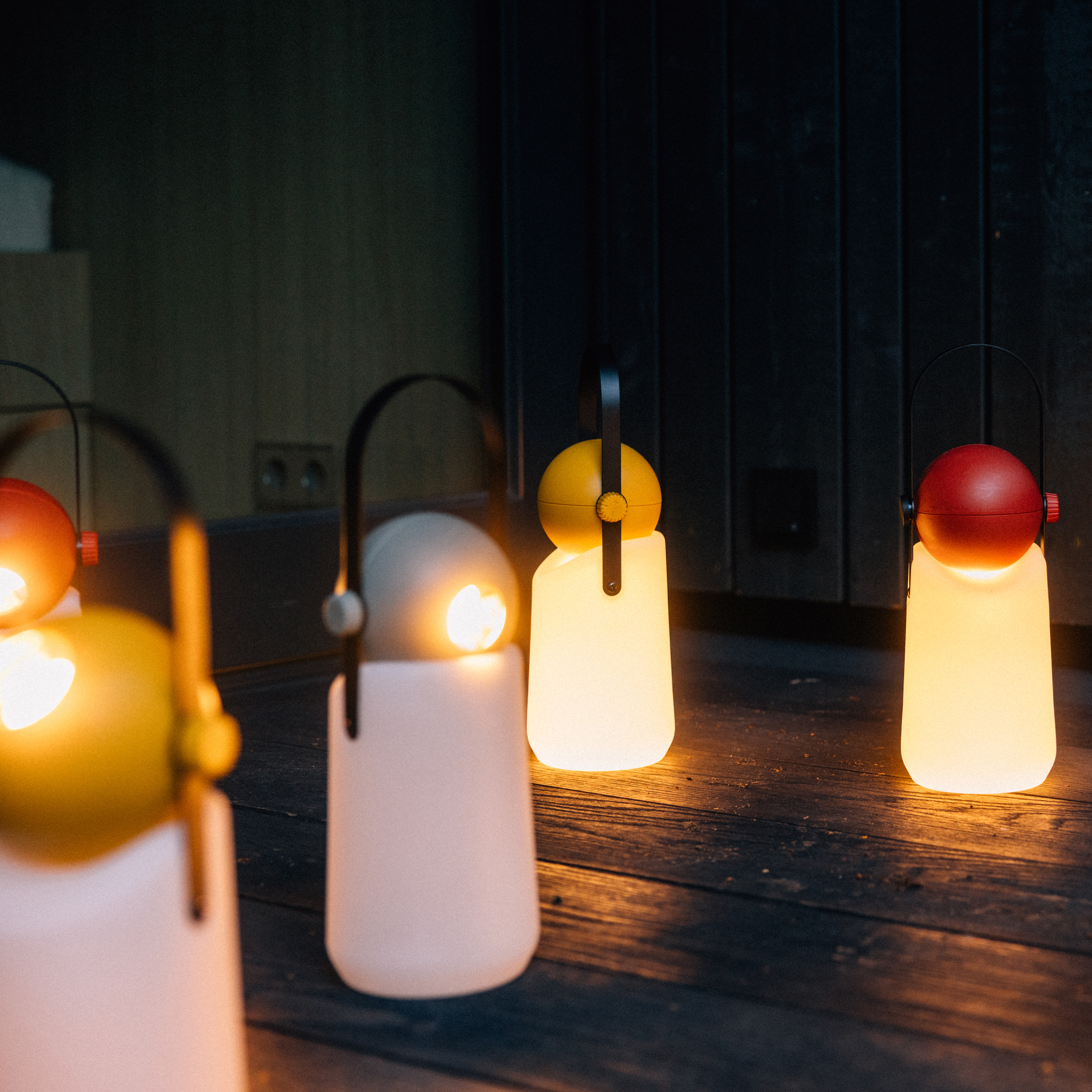 Fünf tragbare Leuchten „Guidelight“ der Marke Weltevree stehen auf einem dunklen Holzboden. Die Leuchten bestehen aus Kunststoff in Weiß mit roten oder gelben Akzenten und erzeugen ein warmweißes Licht. Die Szene wirkt einladend und modern, geeignet für innen und außen.