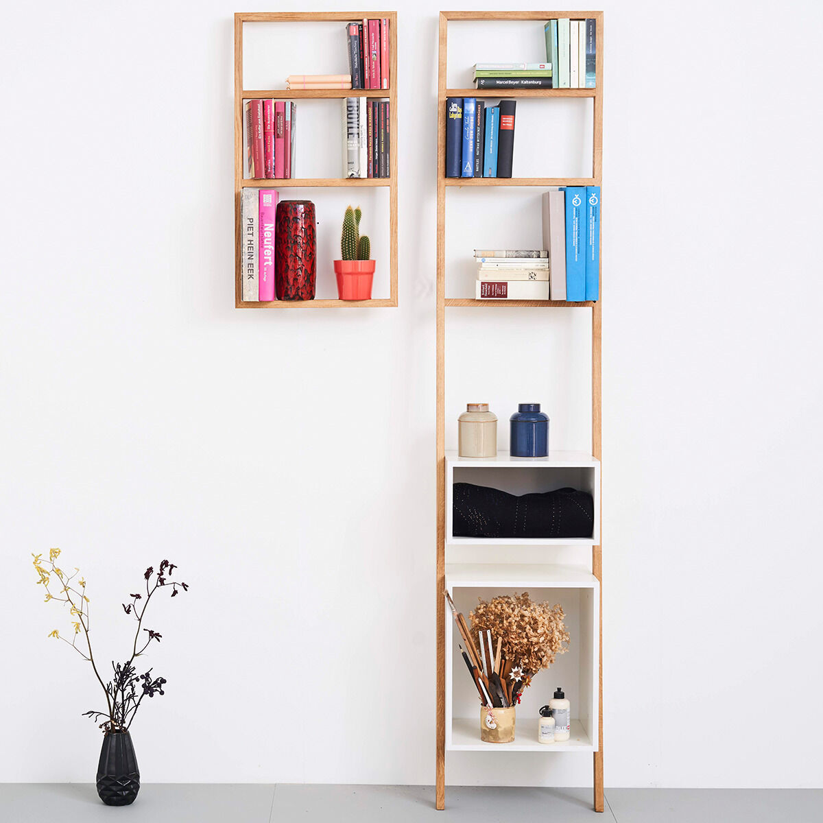 Ein modernes Regal namens »bertolt« aus Holz und weißem Material, das vertikal an einer Wand montiert ist und verschiedene Bücher und dekorative Objekte wie Vasen und eine Kaktee enthält.