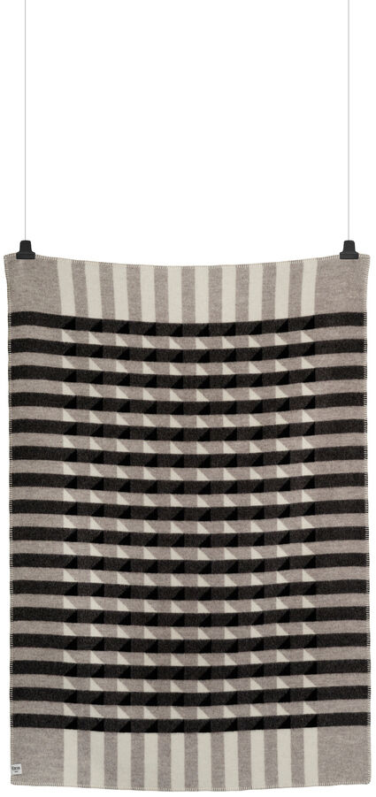 Eine graue, schwarze und beige gestreifte Lammwolldecke namens »KVAM«, die an zwei schwarzen Haken hängt.