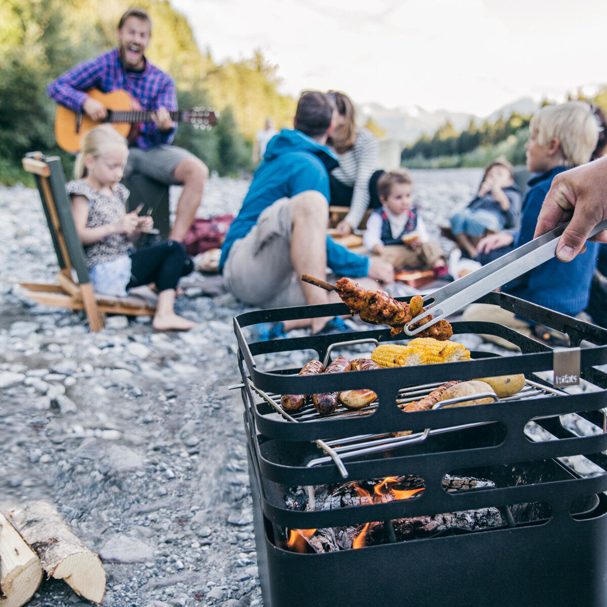 Eine Gruppe von Menschen genießt ein Grillfest im Freien in einer natürlichen Umgebung am Flussufer. Im Vordergrund steht ein tragbarer Holzkohlegrill mit verschiedenen Fleischspießen und Grillgut, während im Hintergrund ein Mann mit Gitarre für musikalische Unterhaltung sorgt. Kinder und Erwachsene sitzen auf dem Boden und auf Campingstühlen und genießen die gemeinsame Zeit.