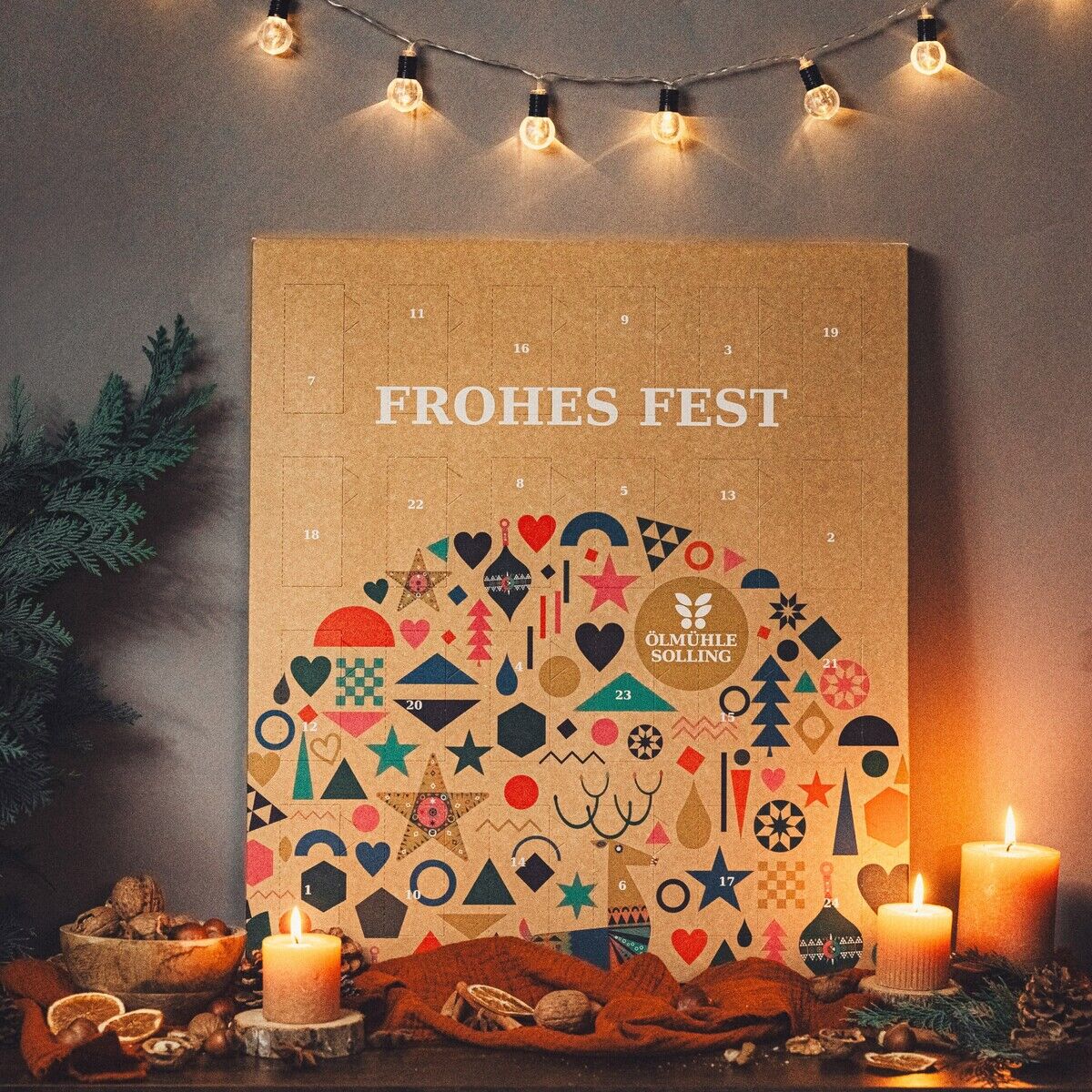 Adventskalender mit dem Schriftzug 'Frohes Fest' steht dekorativ auf einem Tisch, umgeben von brennenden Kerzen, weihnachtlicher Dekoration, getrockneten Orangenscheiben und Nüssen. Im Hintergrund sind Lichterkette und ein grüner Tannenzweig zu sehen. Die Szene vermittelt eine gemütliche, festliche Stimmung.