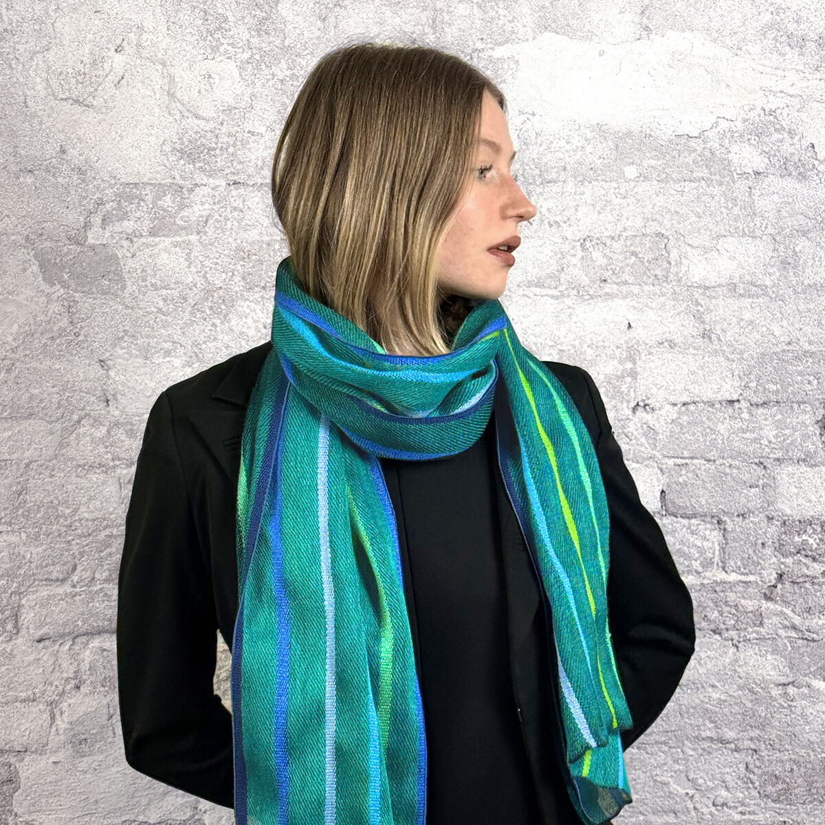 Ein fein gewebter Seidenschal der Marke Ulrike Isensee ist mittig drapiert dargestellt. Das Modell zeigt abwechselnd schmale und breite Querstreifen in kräftigem Blau, weichen Grüntönen, Türkis, Weiß und dezentem Grau. Der Schal hat einen sanften Glanz, wirkt leicht und fällt weich, perfekt als stilvolles Modeaccessoire.