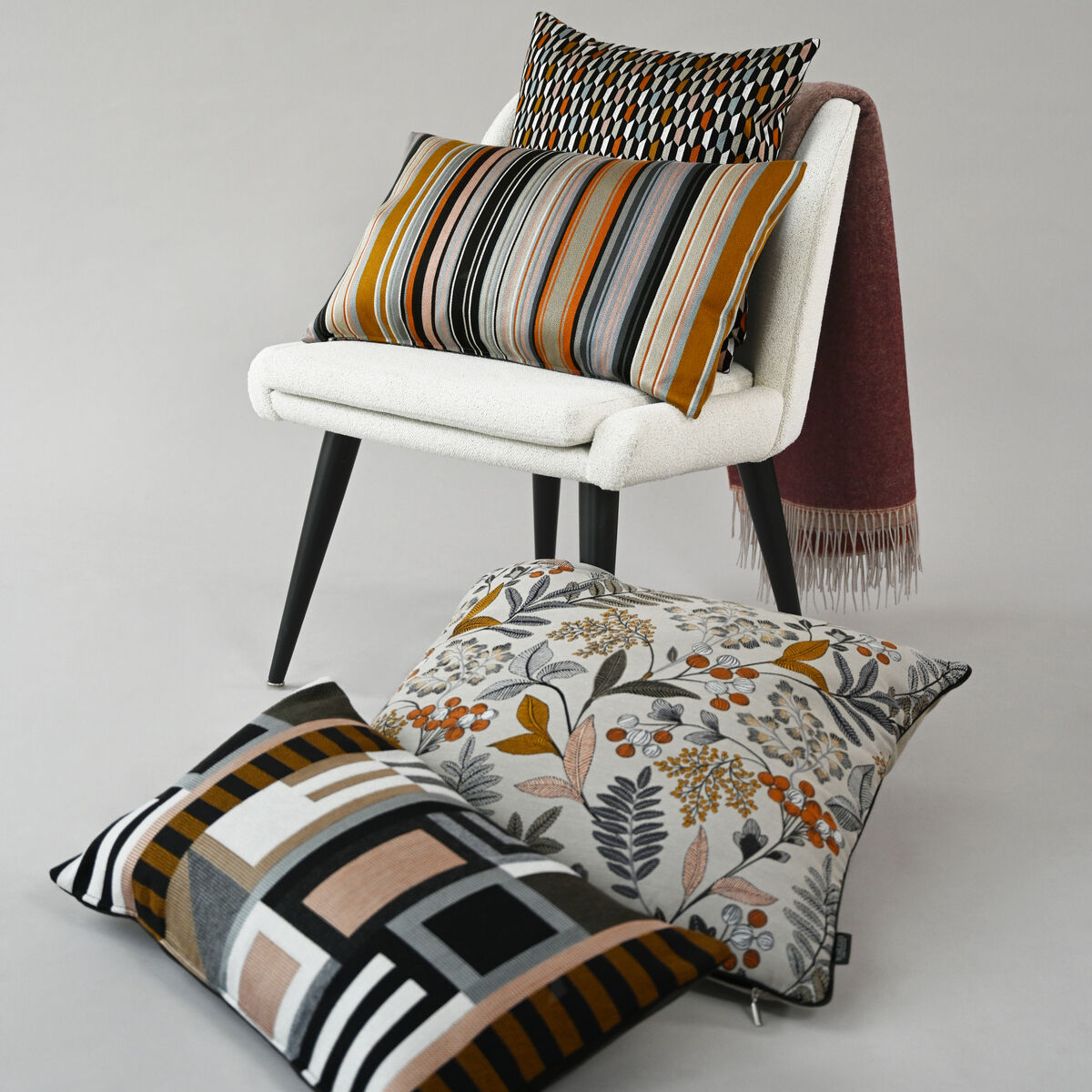 Ein Arrangement von Kissen auf einem weißen Stuhl vor einem neutralen Hintergrund. Die Kissen haben verschiedene Muster, darunter Streifen und florale Designs in Herbstfarben wie Orange, Braun und Grün.